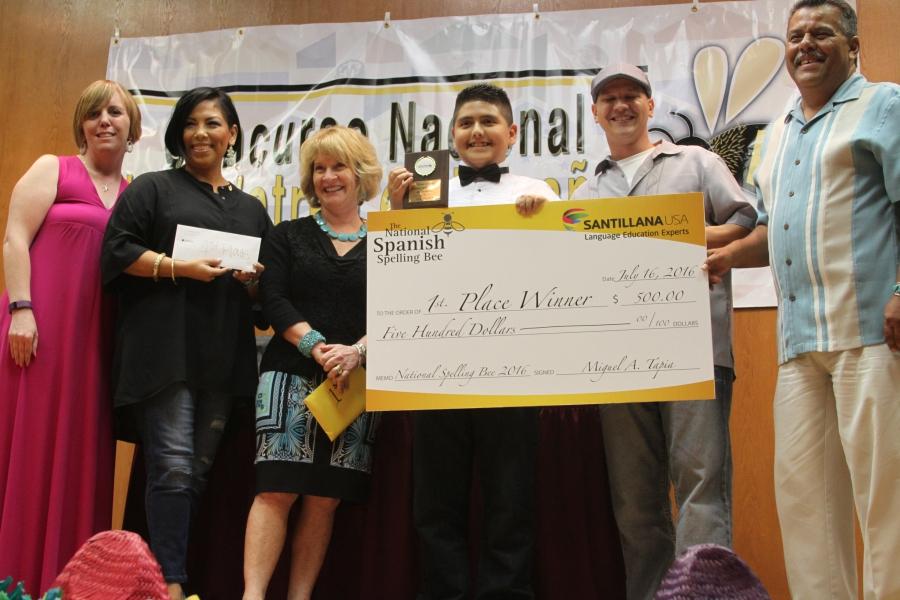 Row of people in front of spelling bee banner, with winner holding giant check and plaque