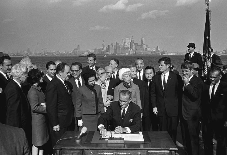 President Lyndon B. Johnson sits at a table and signs the Immigration and Nationality Act on Liberty Island. Vice President Hubert Humphrey, Lady Bird Johnson, Muriel Humphrey, Sen. Edward (Ted) Kennedy, Sen. Robert F. Kennedy, and others look on.
