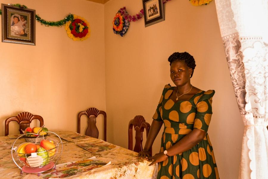 Lika, 29, stands at a dining room table in her home in northern Uganda. 
