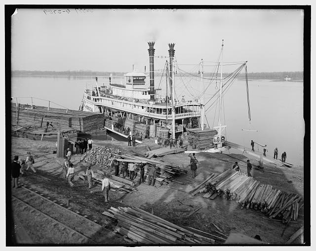 A historic photograph of prisoners at work between 1900 and 1910, carrying lumber from a dock onto a ship in the Mississippi River near Angola prison. 