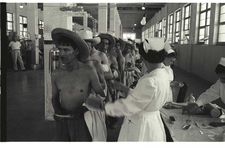 Men without shirts and wide-brimmed hats line up in front of a nurses' table.
