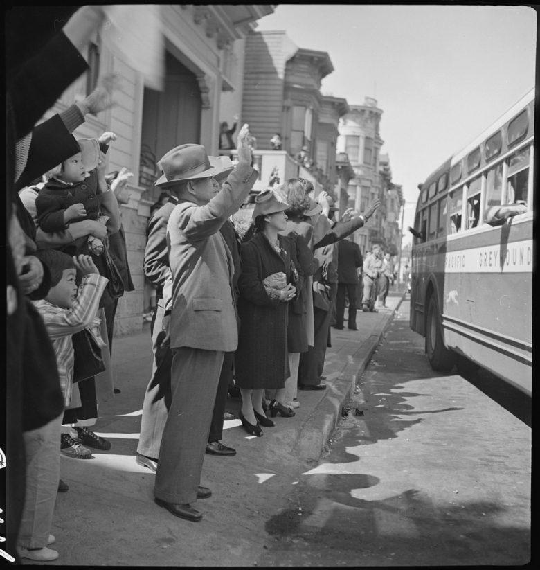 Men and women stand on a sidewalk, waving at a departing Greyhound bus