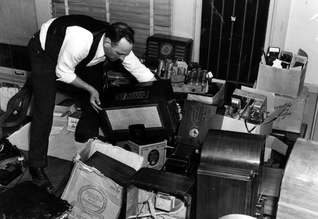 Black and white image of man go through a pile of cameras and radios