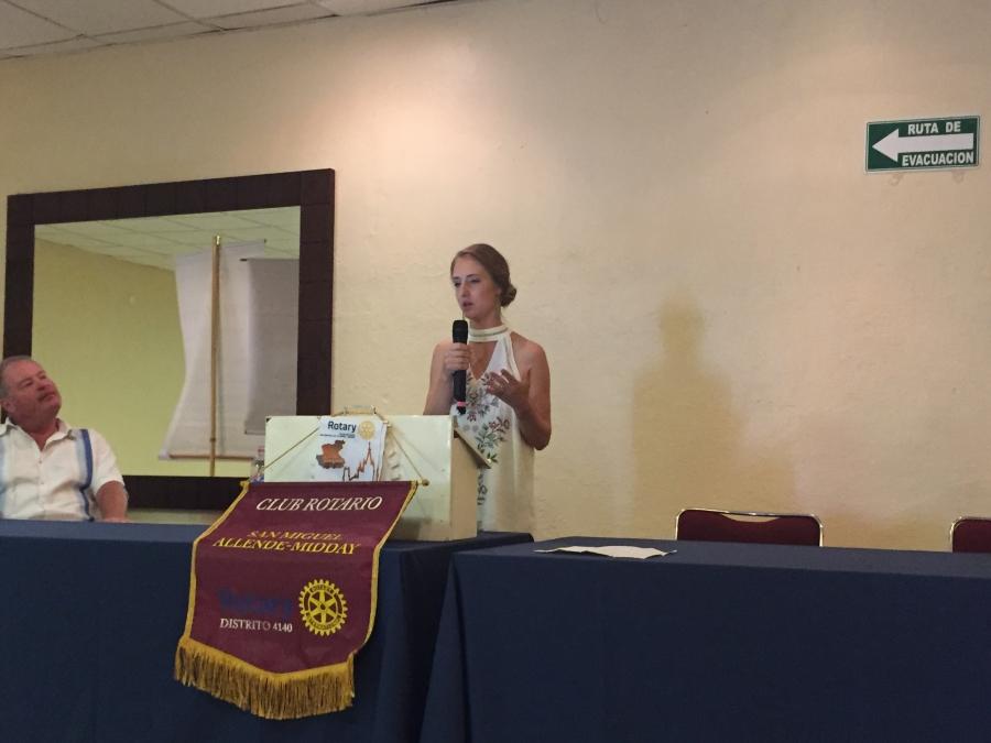 Woman in white dress stands behind podium Rotary Club emblem, holding microphone