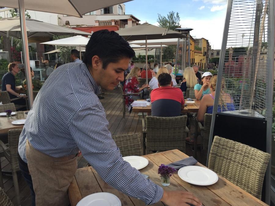 Man setting dishes on table in outdoor restaurant, with customers at table behind