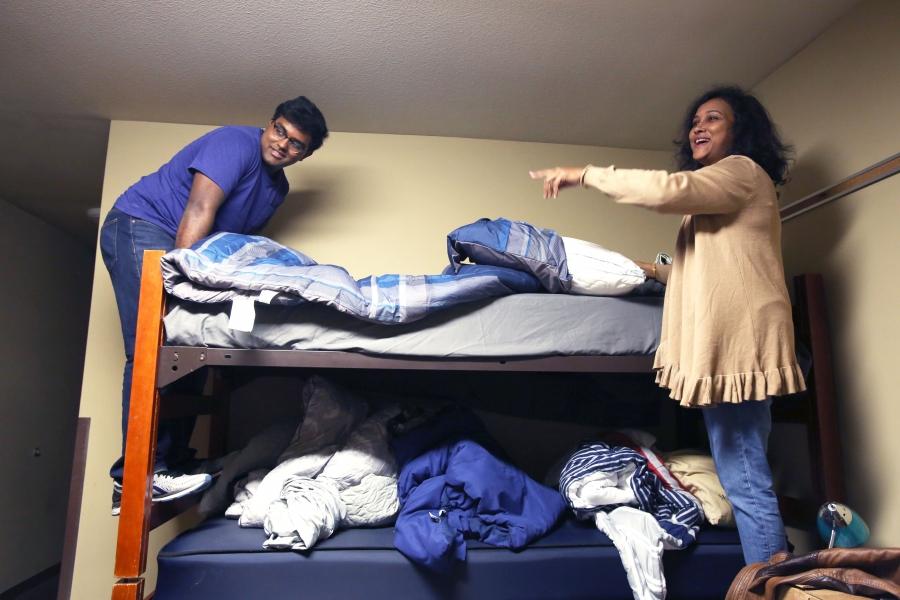 Bunk bed in dorm room, young man and woman stand on either side of top bunk, making the bed