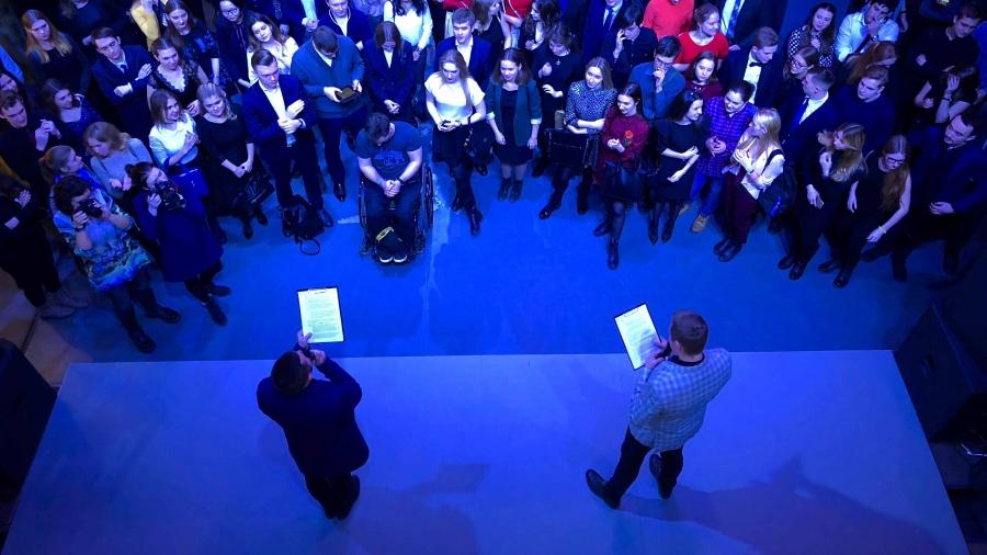 The view overhead of a crowd gathered around a stage while two men read from clipboards. The room is bathed in blue light. 
