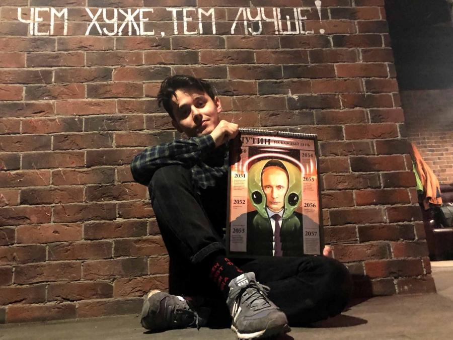 A young man poses with an oversized calendar against a brick wall. The cover of the calendar has an illustration of Vladimir Putin wearing an alien-shaped helmet.