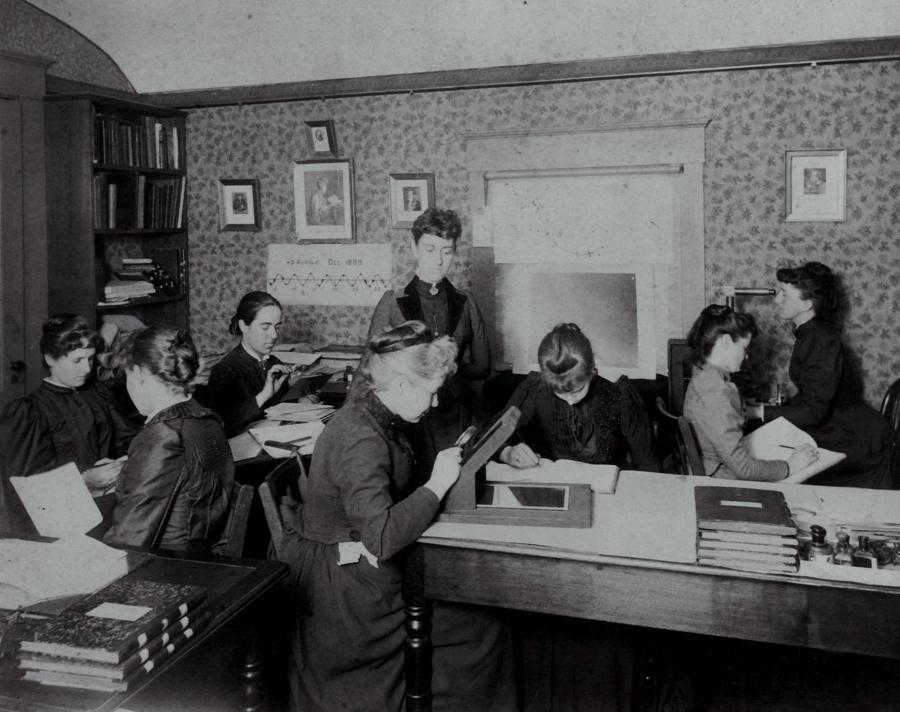 A group of women are sitting in a tiny room, some holding magnifying glasses, bent over boxes studying the glass plates. One stands in the center of the room, overseeing them. This is Williamina Fleming. 