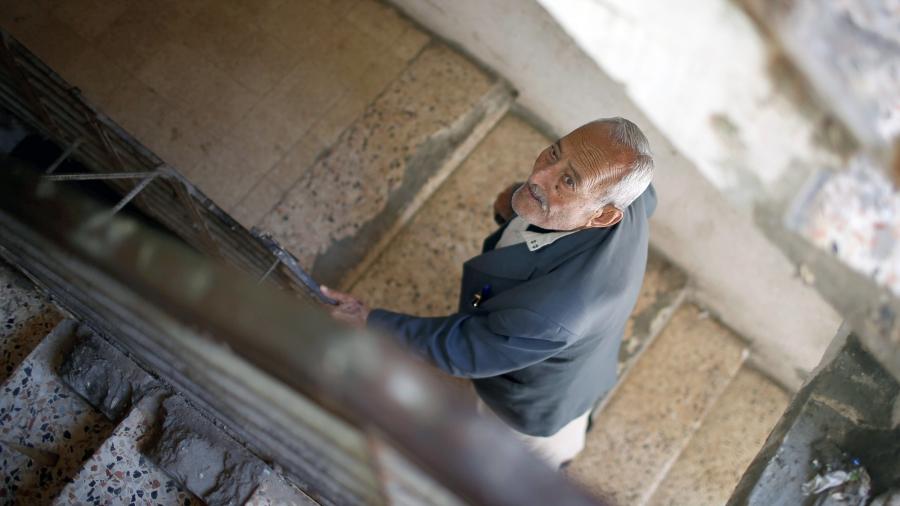 An older man looks up as he hangs onto the bannister as he climbs stairs in a home.