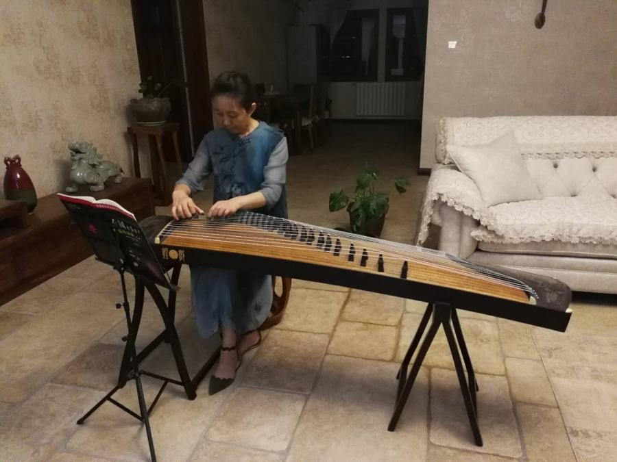 A woman in a blue dress sits facing the camera and a music stand and plays guzheng, a Chinese traditional instrument that looks like a small table with strings on it
