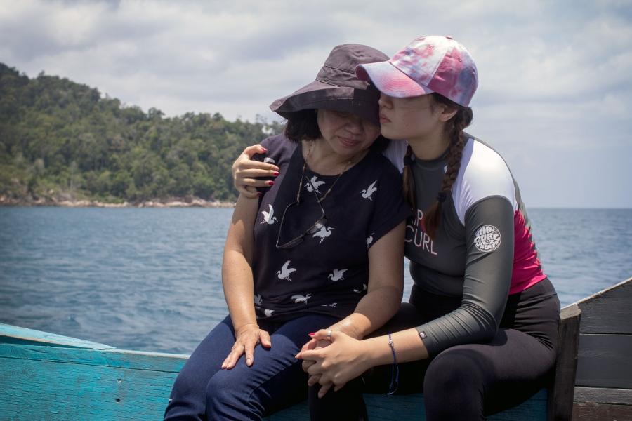 A younger woman wraps her arms around an older woman as they sit on the edge of boat.