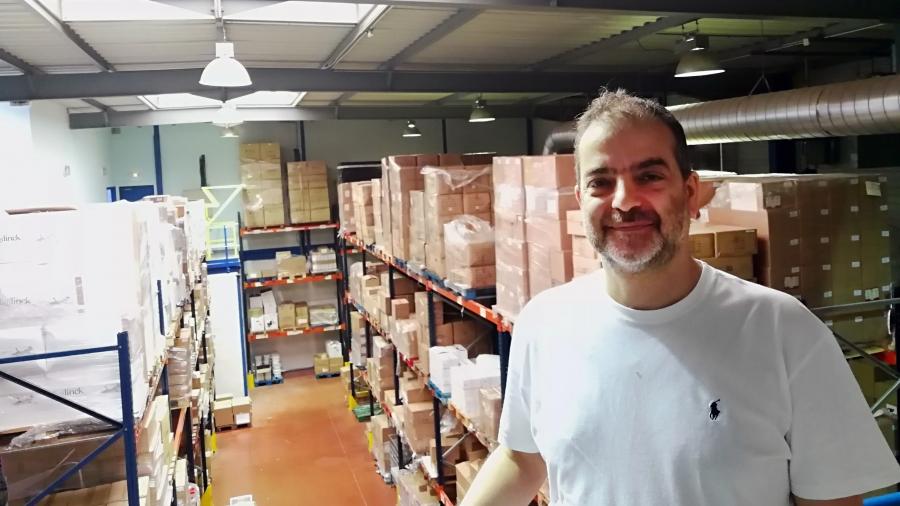 A man stands in a warehouse. Behind him, boxes line the shelves. 