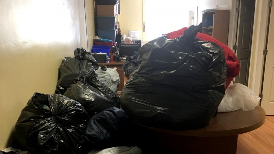 Piles of trash bags are stacked in an office. In the distance, a woman is working on a computer.