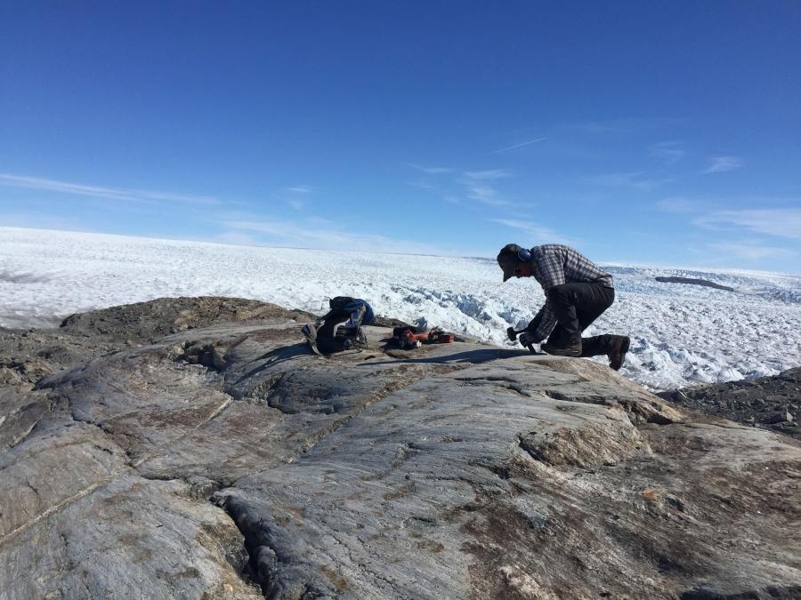 A man kneels on rock at the edge of the Greenland ice sheet.