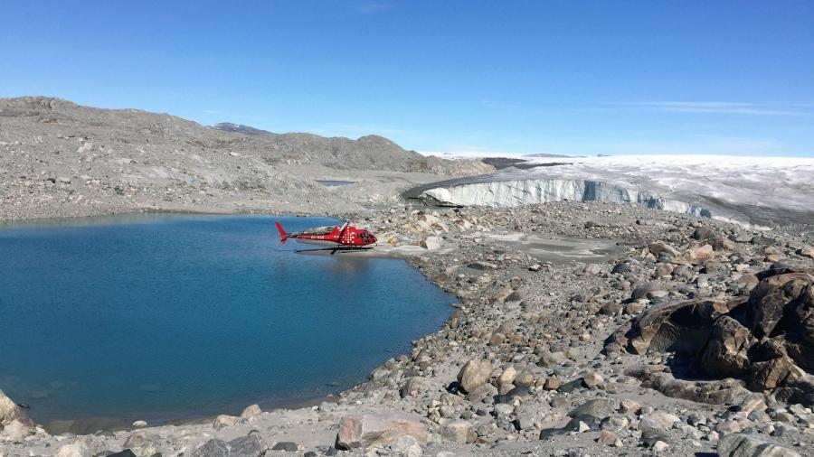 A red helicopter is landed on the edge of a blue lake on a rocky shoreline. Hills in the distance are covered in white ice. 