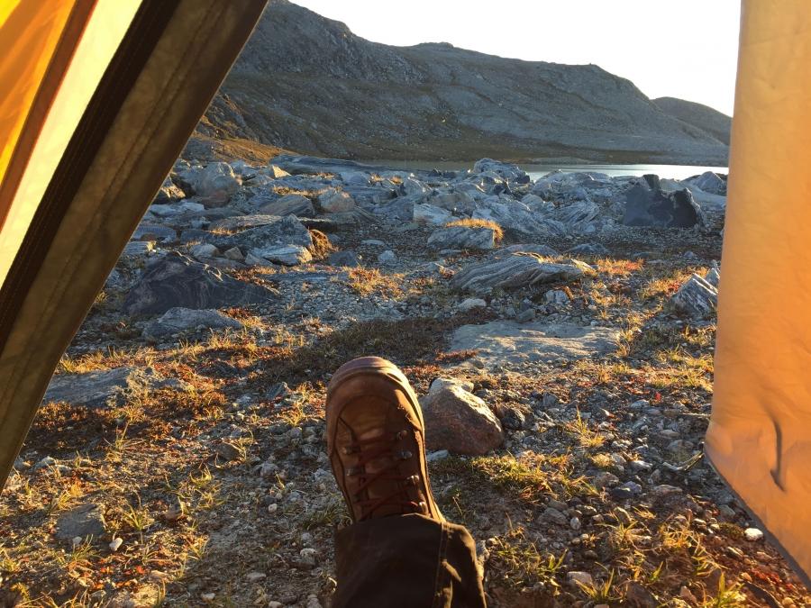 A woman's booted foot is framed by the walls of her tent as the golden sunlight spills over rocky terrain. 