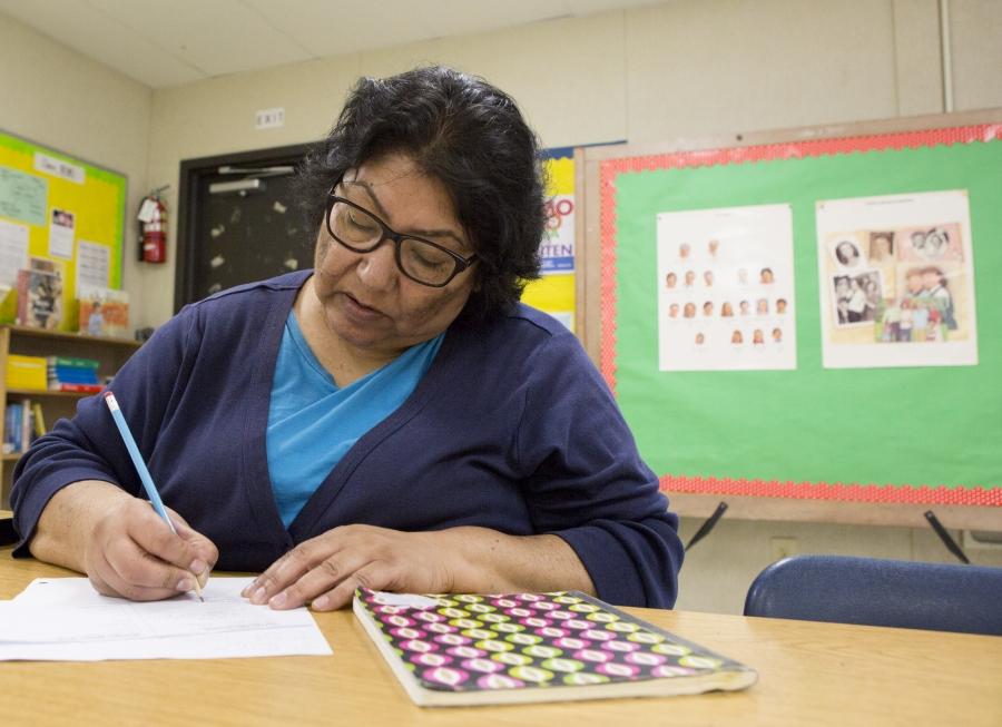 Woman sits at desk in elementary classroom, writing on sheet of paper with a pen