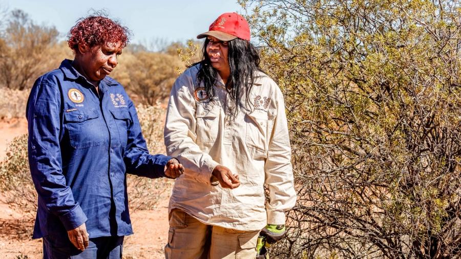 Two women speak to each other as they walk through the bush in an arid desert.