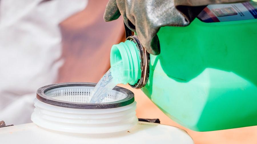 A gloved hand holds a green bottle and pours a clear liquid into another plastic container.