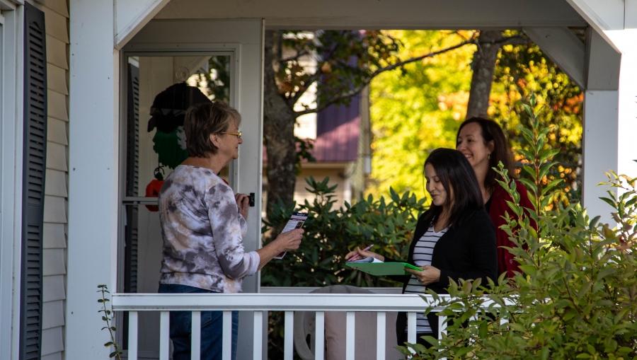 Safiya Wazir is shown with a green clipboard in hand standing on the front porch, speaking with a resident of Concord, New Hampshire.