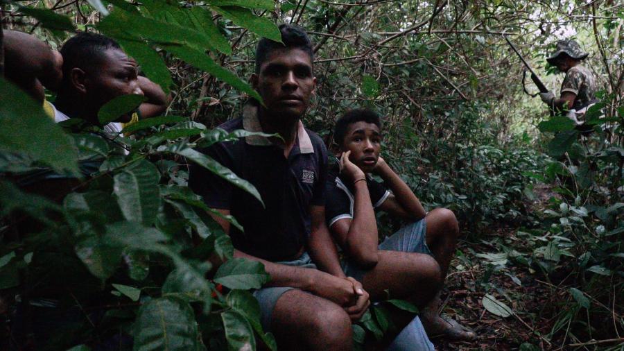 Three boys are on their knees in the jungle, with their hands clasped behind their heads.