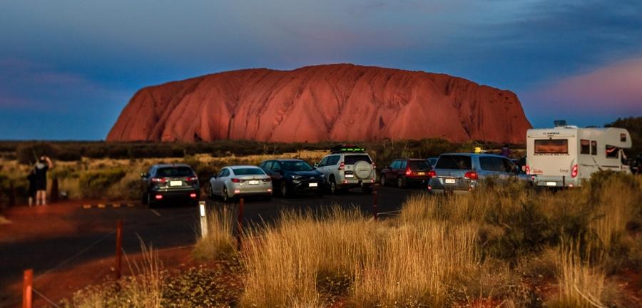 Cars are shown in the foreground near the base of Uluru, cast in red light from the sunset.