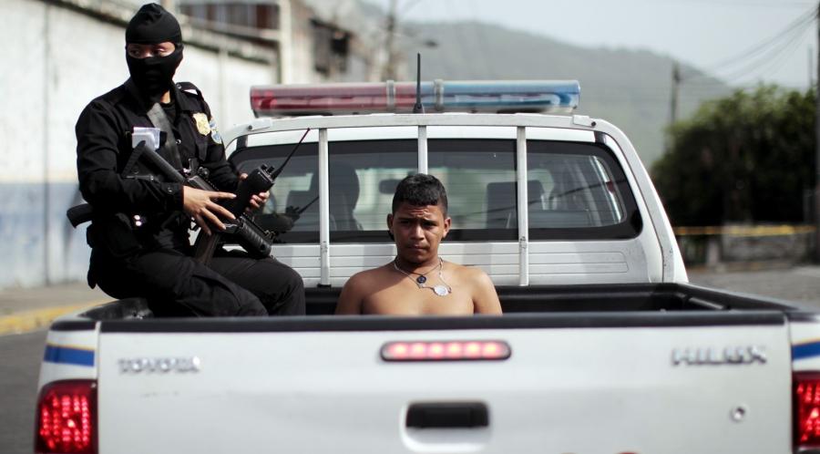 Policewoman in black mask with gun detains shirtless youth sitting in back of police pick-up truck. 