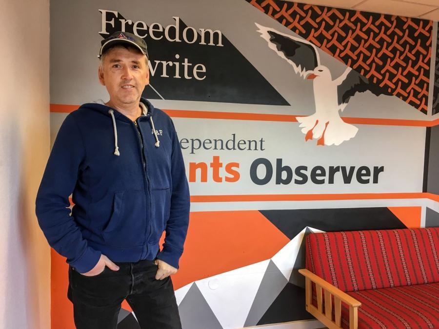 A man stands for a portrait in front of a painted wall that says Barents Observer