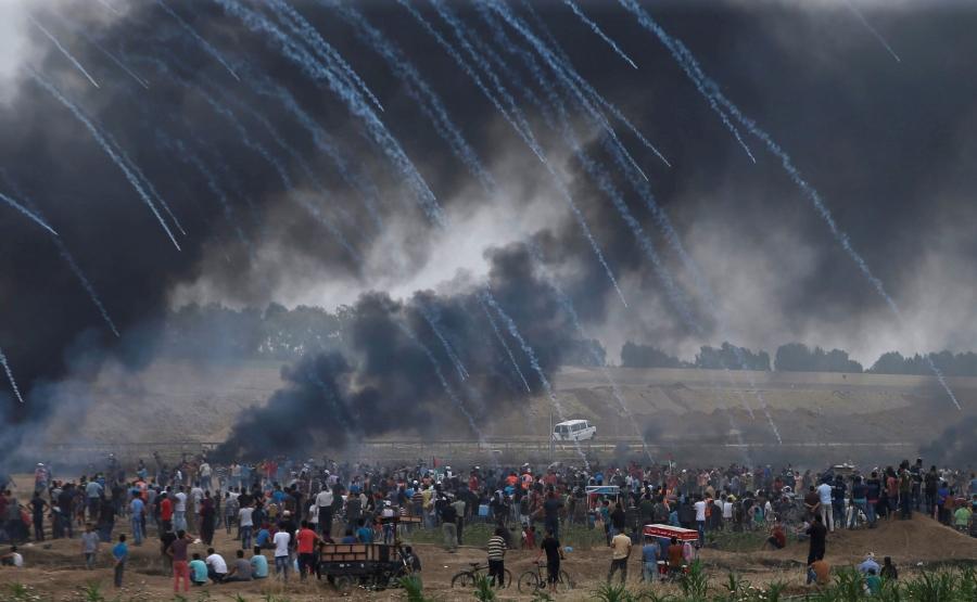 A crowd of people stand in a group. Overhead, gas trails behind tear gas canisters as they're launched into the crowd.