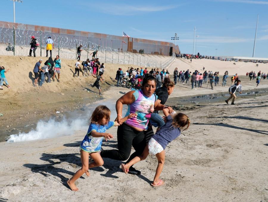 A woman holds the hands of two children as she runs away from the US-Mexico border fence. Behind her are more people running and clouds of tear gas.