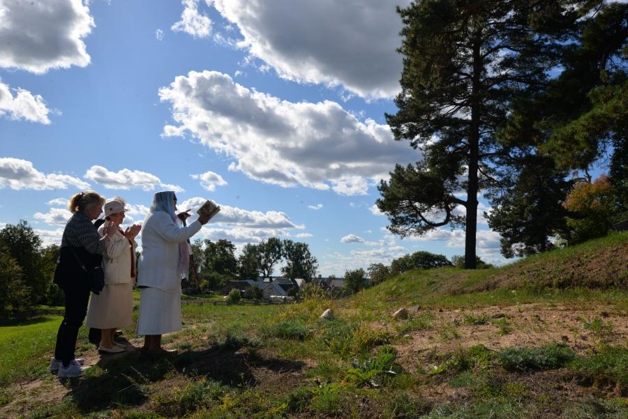 Three women stand green hill, facing gravestone