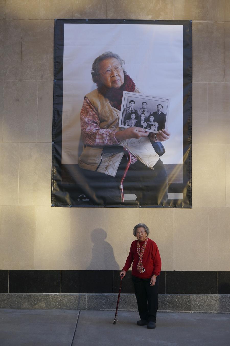 A woman stands in front of her portrait which hangs large on the side of a building