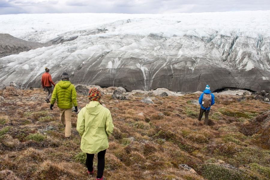 People walk up a hill to the edge of the Greenland ice sheet, which is ice and show as far as the ice can see along the horizon.