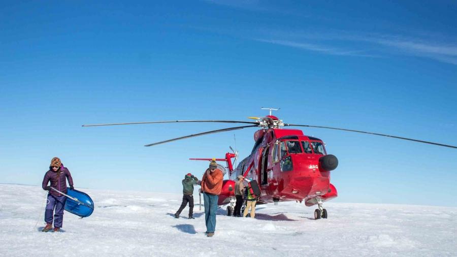 A bright red helicopter lands on the flat ice and snow while people help unload gear from it. 