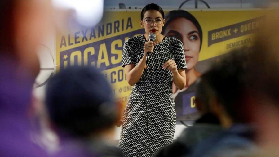Alexandria Ocasio-Cortez holds a microphone in front of a large campaign banner showing her face