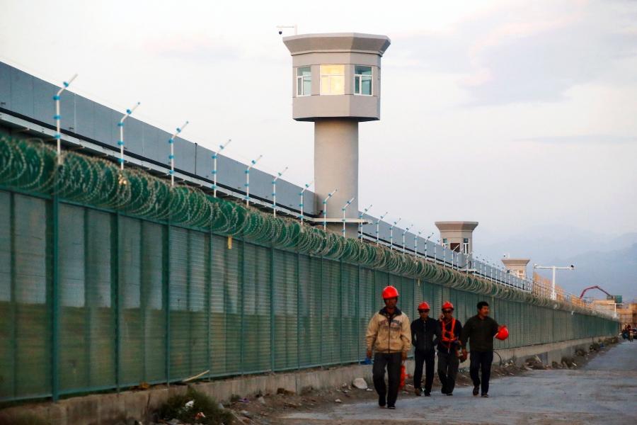 Men in orange hard hats walk alongside a large fence topped with barbed wire. Behind the fence are lookout towers.