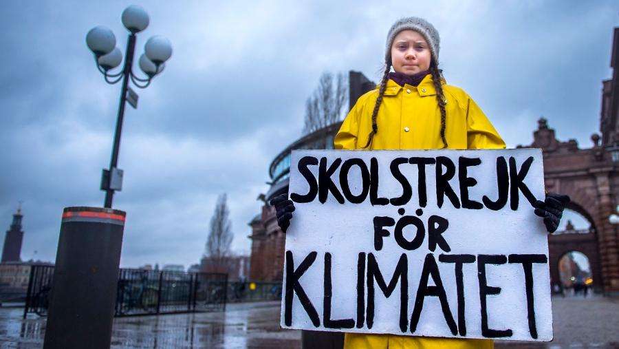 A young girl stands in front of a government building holding a sign in Swedish. Translated, it says: "School strike for the climate"