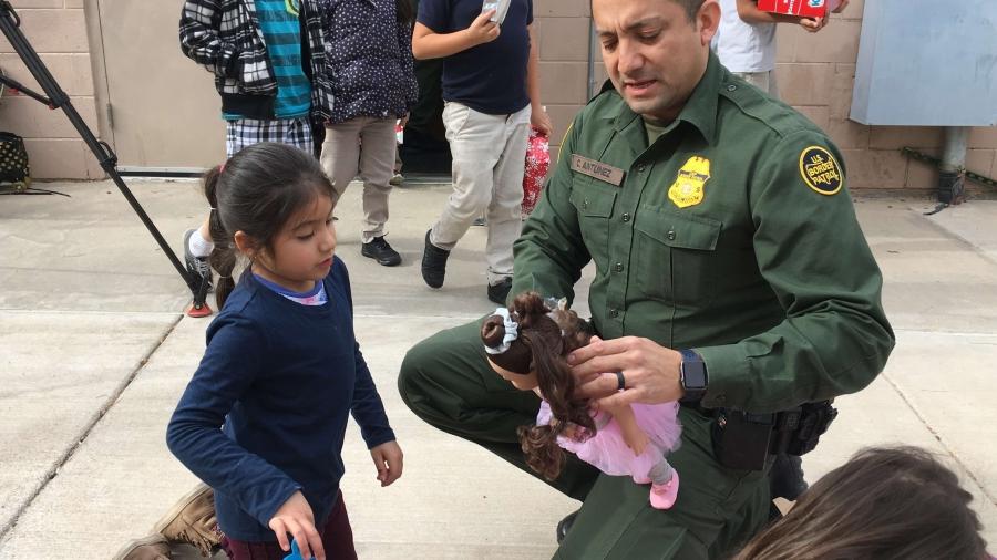 Border patrol agent helps a young girl dress a doll. 