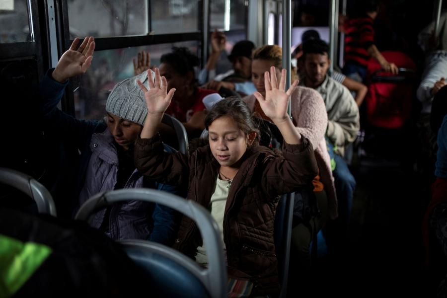 Migrants are shown with their hands raised in the air praying on a bus.