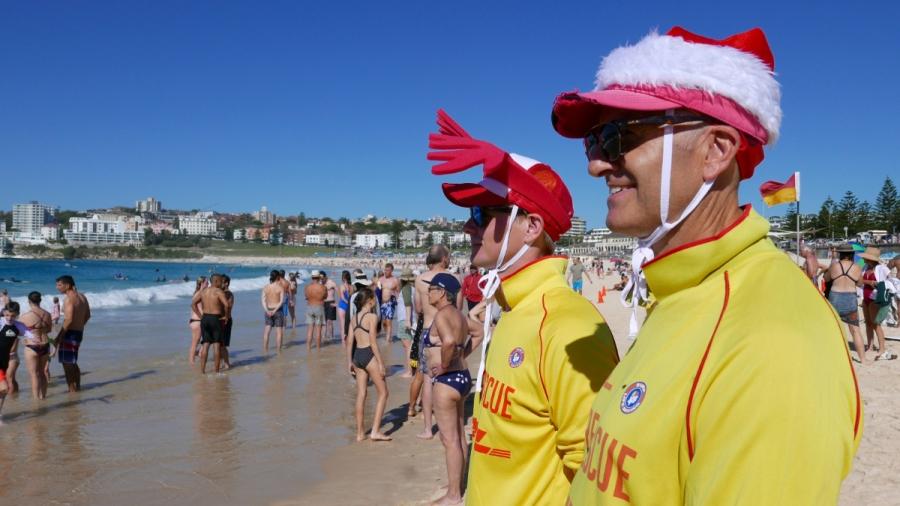 Two men wearing Santa Claus's hats stand on a sunny beach surrounded by people in bathing suits in Sidney Australia.