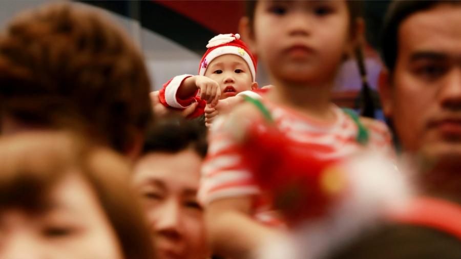 A toddler is dressed up as Santa Claus and looks at the camera while looking over a crowd of people.