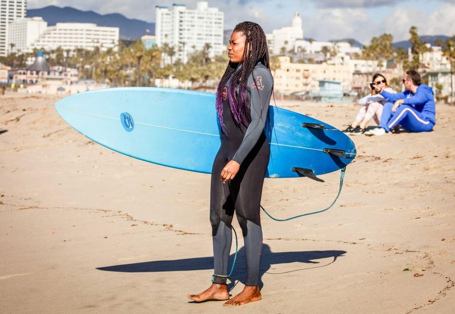 A woman stands on a beach carrying a surfboard under her arm. 