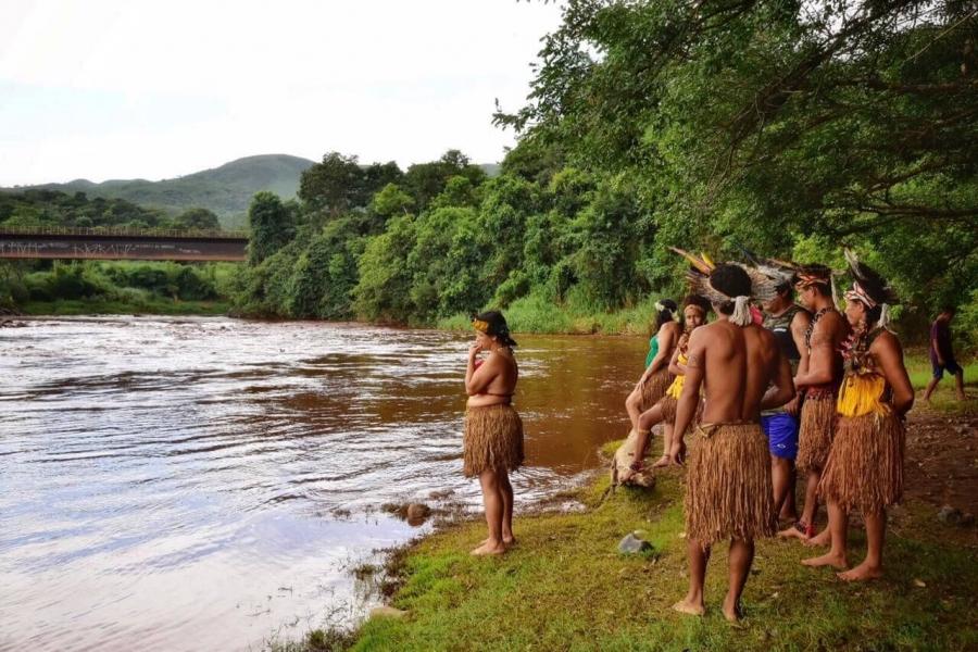 Indigenous people stand on the banks of a flooded river 