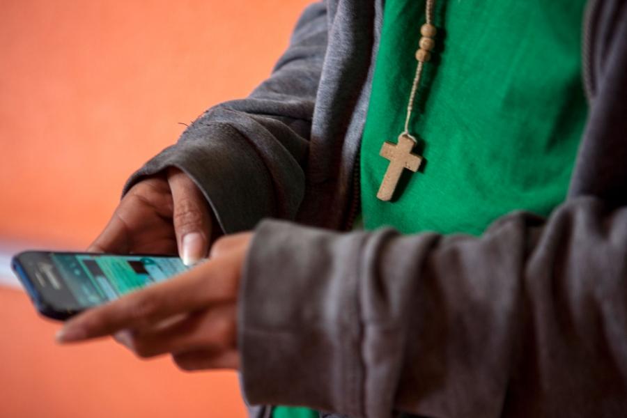 A teenage boy holds a cell phone in his hands. A wooden cross hanging from his neck dangles into the frame. His face is not pictured. 
