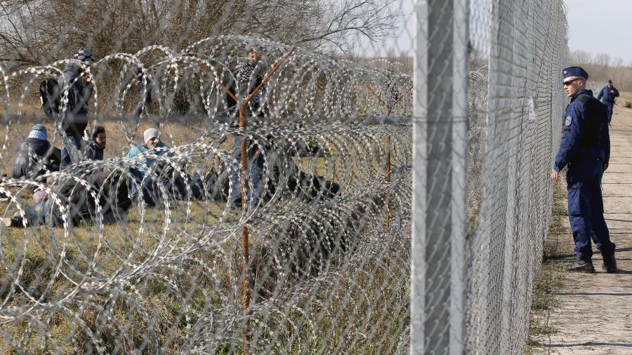 A Hungarian police officer, right, watches migrants through a chain link fence and a fence made of razor wire.