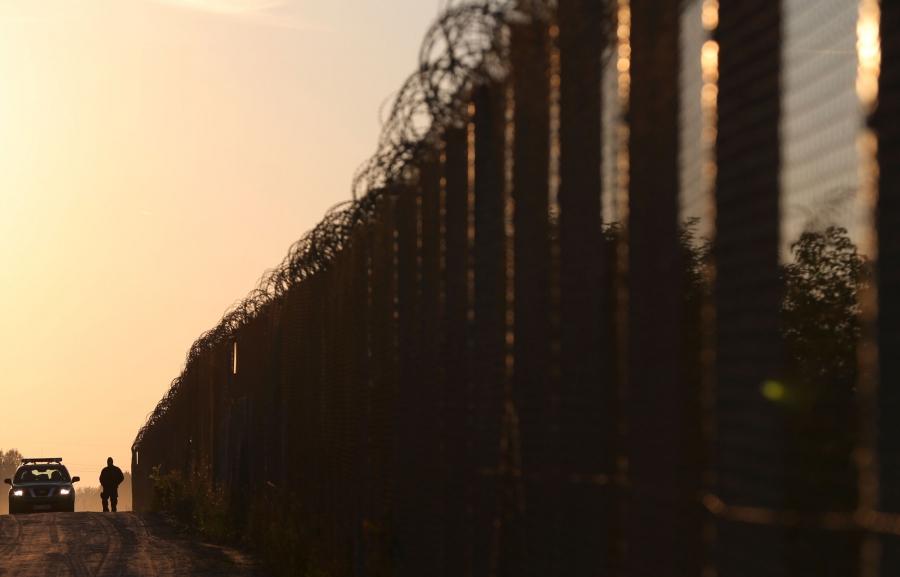 A police officer and a tall fence topped with razor wire is silhouetted against an orange sky. 