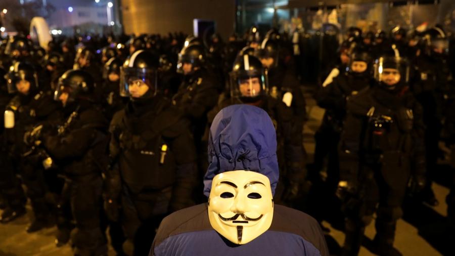 A man stands with his back to the camera. On the back of his head is a Guy Fawkes mask. In front of him are dozens of police wearing riot gear. 