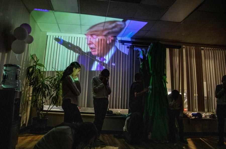 Images of Donald Trump is projected on the wall of a building. Silhouettes of the actors are in front of the image.