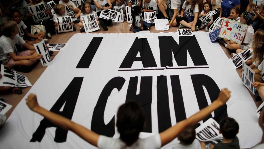 Young children hold signs that say "I am a child" and sit around a large banner that says "I am a child"
